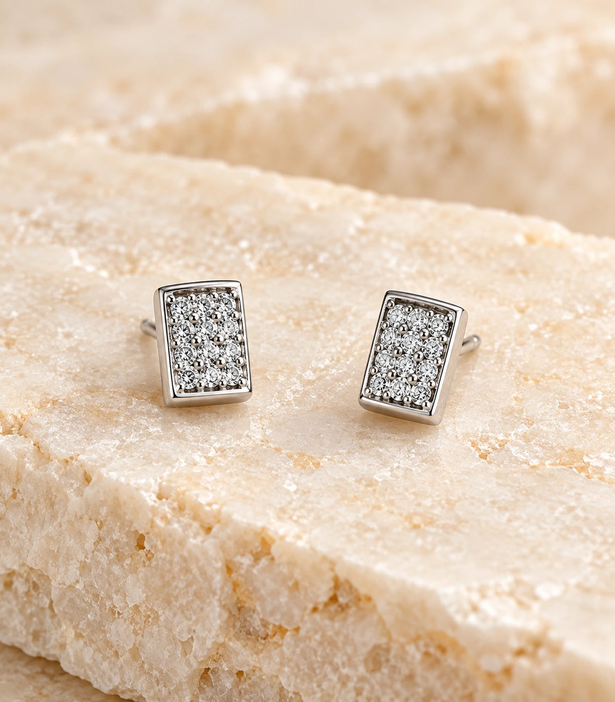 Close view of the pair of silver-tone rectangular pavé stud earrings on beige stone