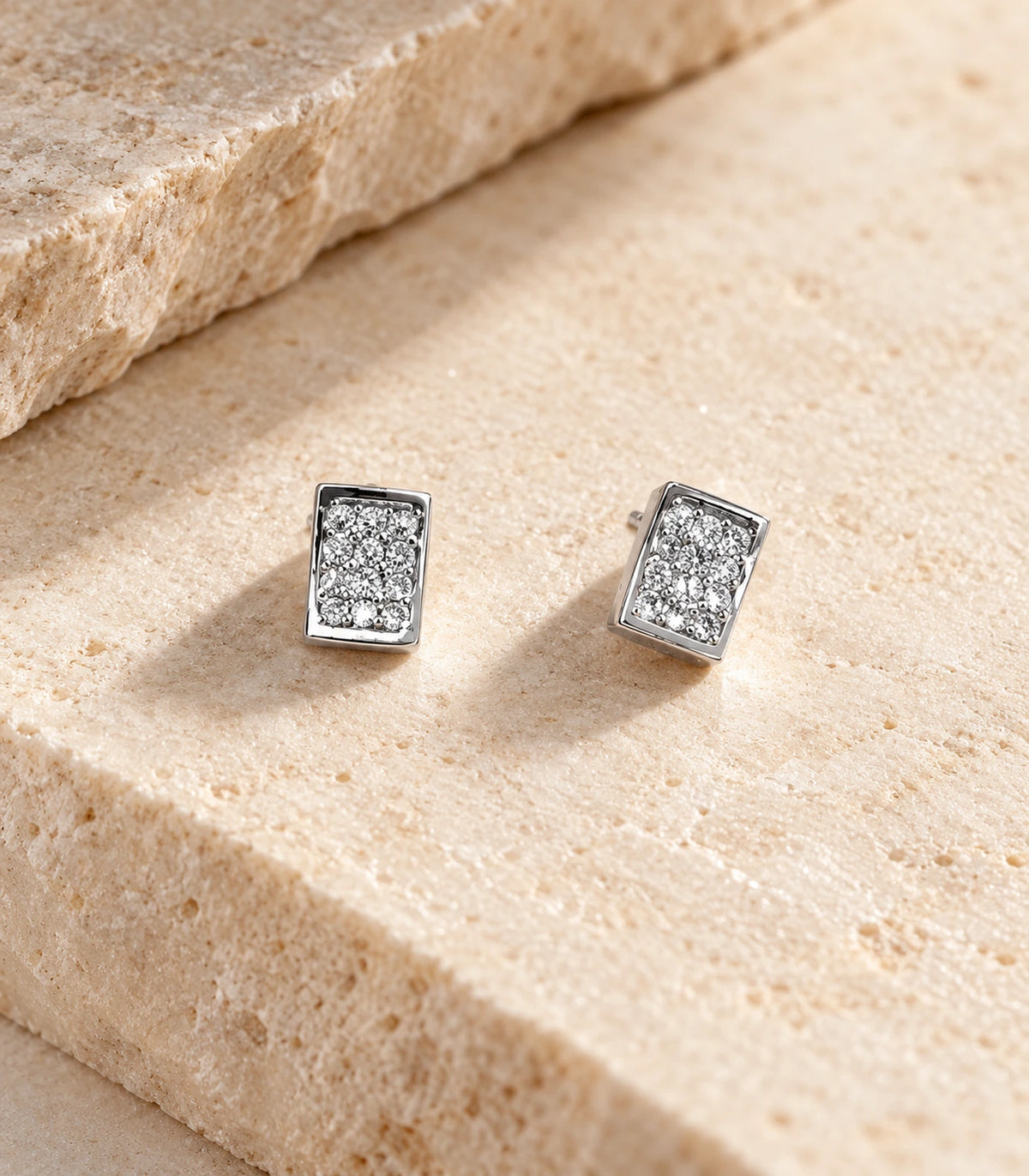 Pair of silver-tone rectangular pavé stud earrings arranged on a beige stone surface