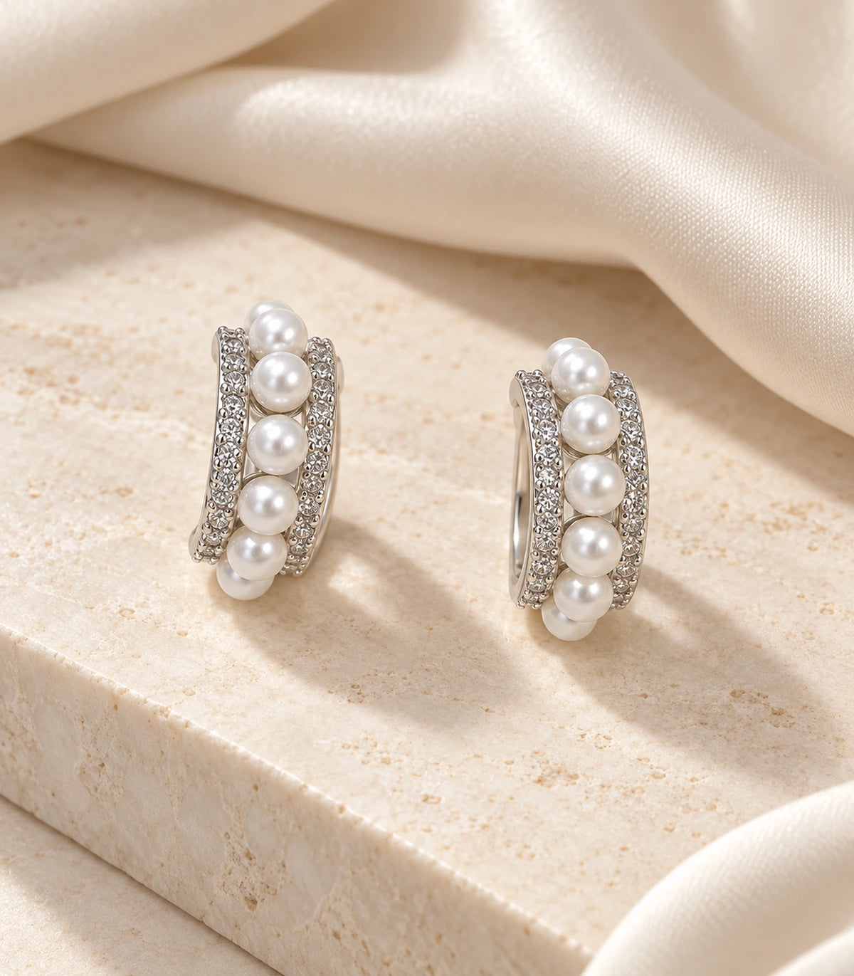 Matching silver-tone pearl and pavé huggie earrings styled on a pale stone surface