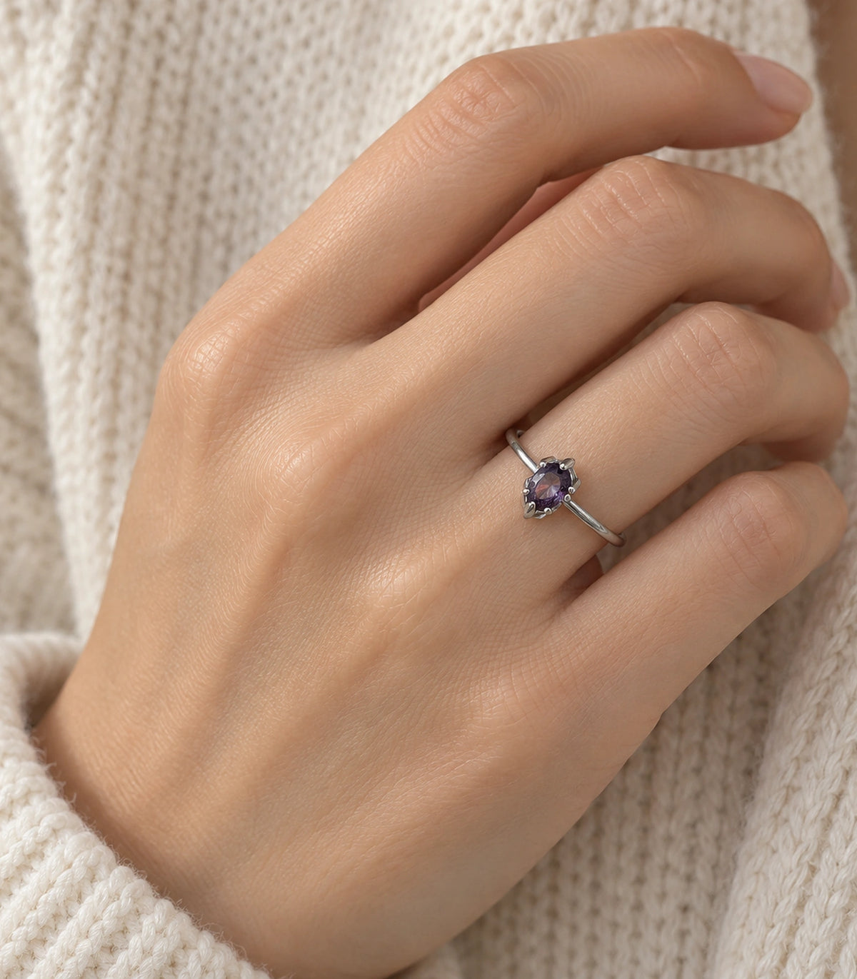 Purple oval solitaire ring worn on a hand against an ivory knit garment