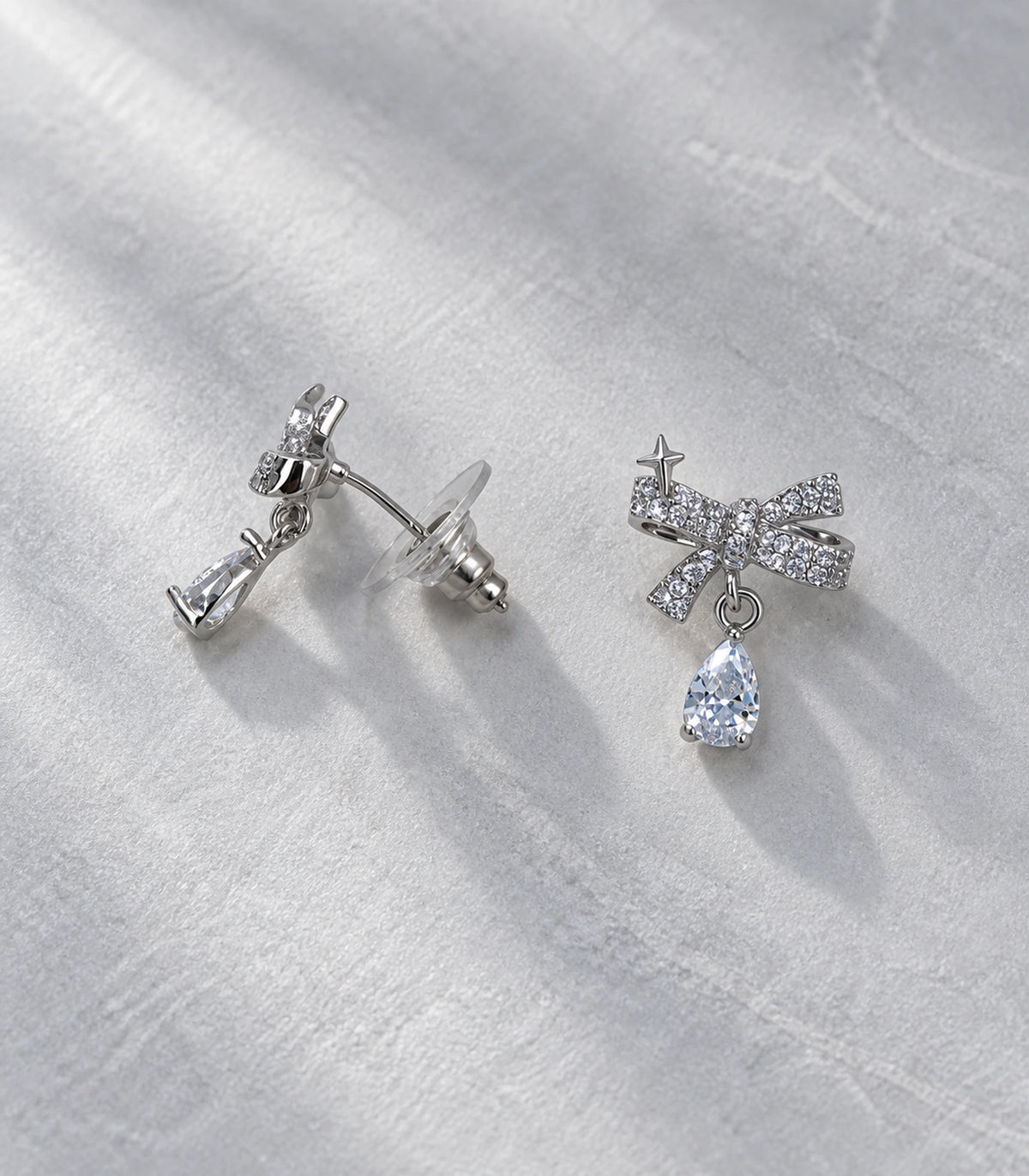 JIMIKI Silver-Tone Star Bow Pear-Drop Earrings in Silver / Clear, editorial flat lay