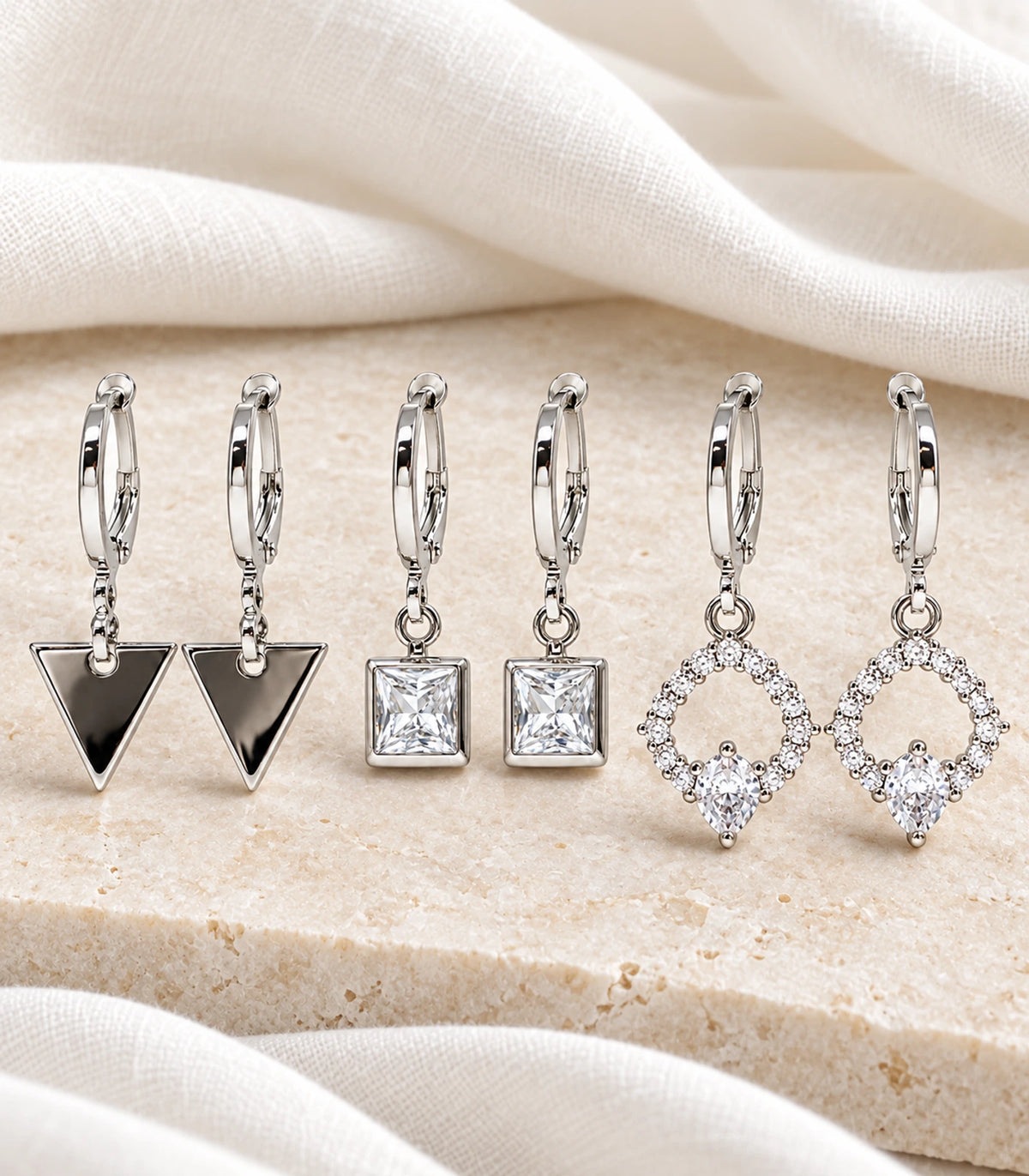 Complete three-pair silver-tone geometric crystal hoop earring set arranged on stone and linen